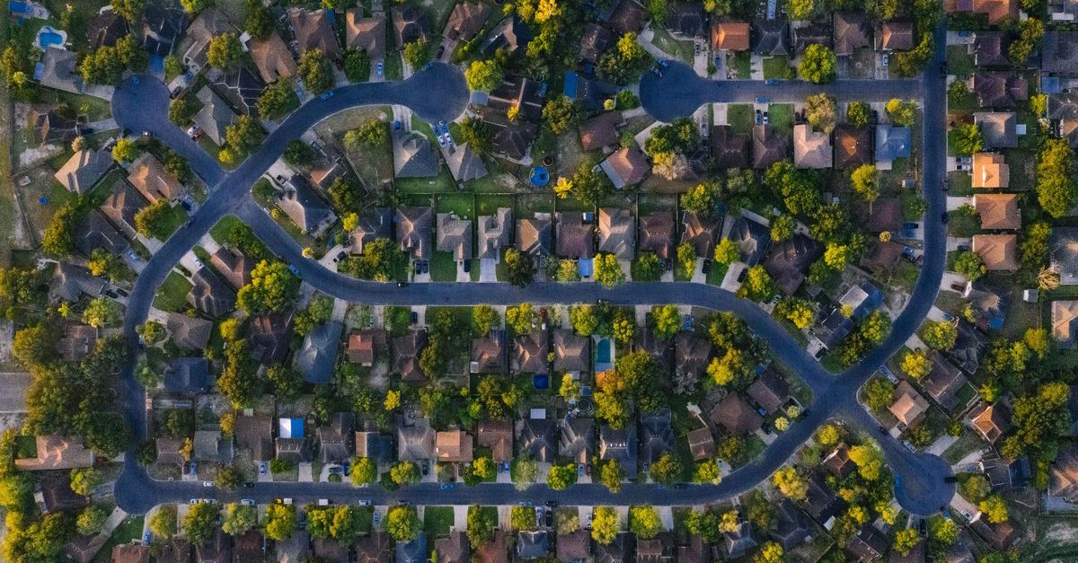 Aerial view of a quiet Texas suburban neighborhood with curving streets, cul-de-sacs, single-family homes with red and tan roofs, mature trees, and late-afternoon light