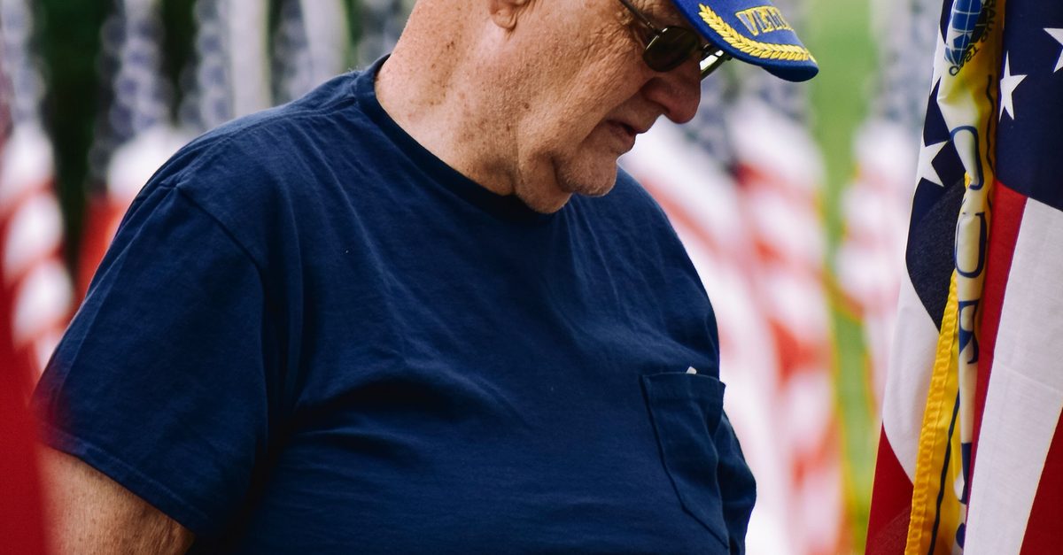 Senior veteran in a blue cap stands beside American flags at an outdoor Texas ceremony